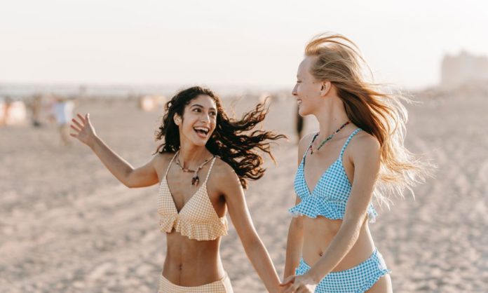 Two women in bikinis - colours are different but the styles are the same - are on a beach. Both look happy as if travel has been really good for them and their mental health