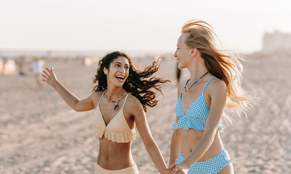You see two women, close friends or besties, out presumably on their bestie-break. One is wearing a yellow checkered bikini and another is wearing a blue checkered bikini. Both are looking at each other and smiling, one has wavy dark brown hair, another has straighter light brown brown hair. 