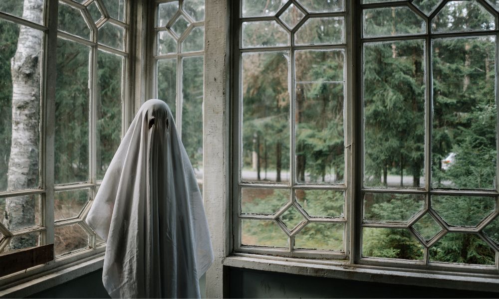 A person in a ghost costume stands inside a room which has full length glass windows and a very spooky feel. The setting looks very haunted  