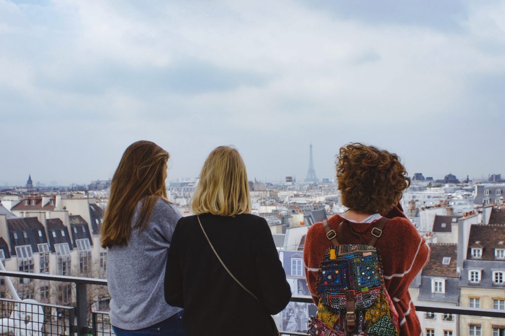 You see three women with their back to the camera looking out from a viewing balcony. You can see the outlines of the city, the unmistakable shape of the Eiffel tower -- Paris is just perfect for a bestie break. One of the woman has a very colourful backpack, patchwork of embroidery and colours.