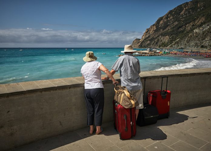 A couple, with their backs to the camera are leaning across a wall and overlooking the ocean and views. There is a suitcase beside them that shows that they are tourists or travellers