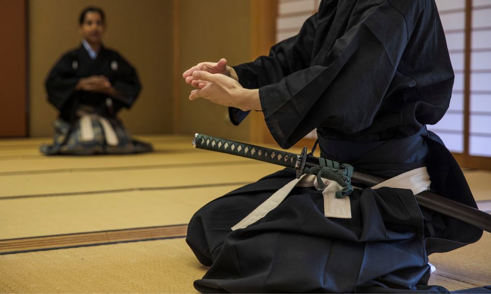 Two samurais in traditional black robes are kneeling down and you can see their swords, though not their faces