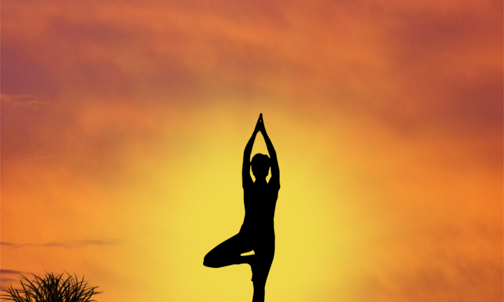 A woman does yoga under an orange and yellow sky - taking time out for wellness and mental health wellbeing while out on a travel. 