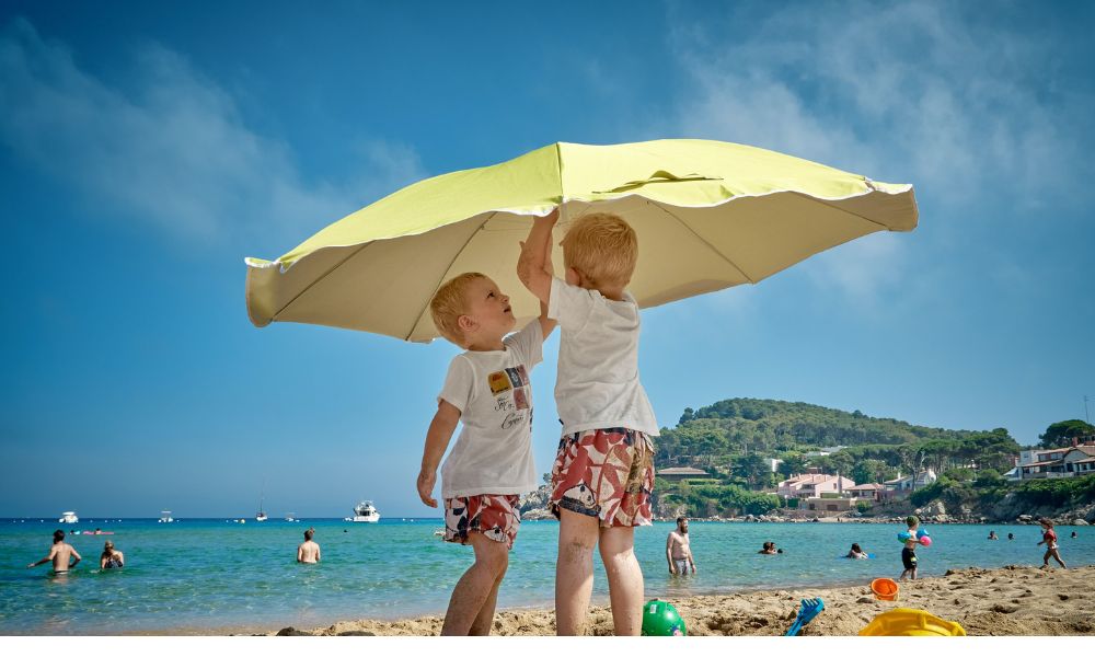 From left to right - two little boys in matching outfits under a beach umbrella. They seem to be brothers, and you can see the sea in the background. In the second photograph, which seems like it could be from Malaga, a woman in a beach outfit walks past other women on both sides. The other women are resting under a canopy of beach huts and in beach wear as well. In the third pic you can see the setting sun as reflected in the waters; the sky is made of various colours of blue. In the last pic, a woman who seems happy, with wind in her hair, and a shy smile holds up her skirt as she dips her feet into the sea, waves at her ankles 