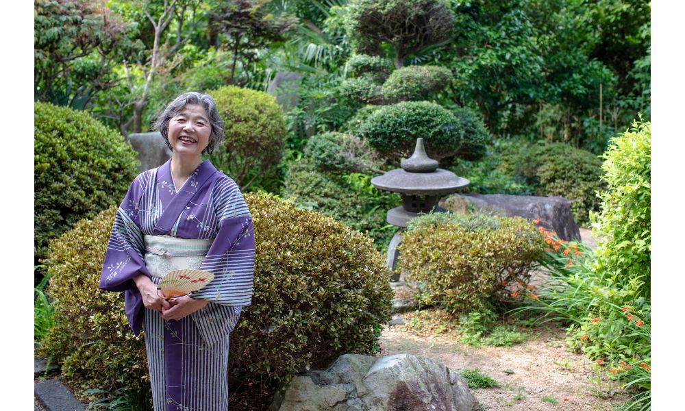 You see a woman in a kimono and a fan standing outside - the background is a beautifully kept together Japanese garden. The woman is smiling and has sliver short hair. 