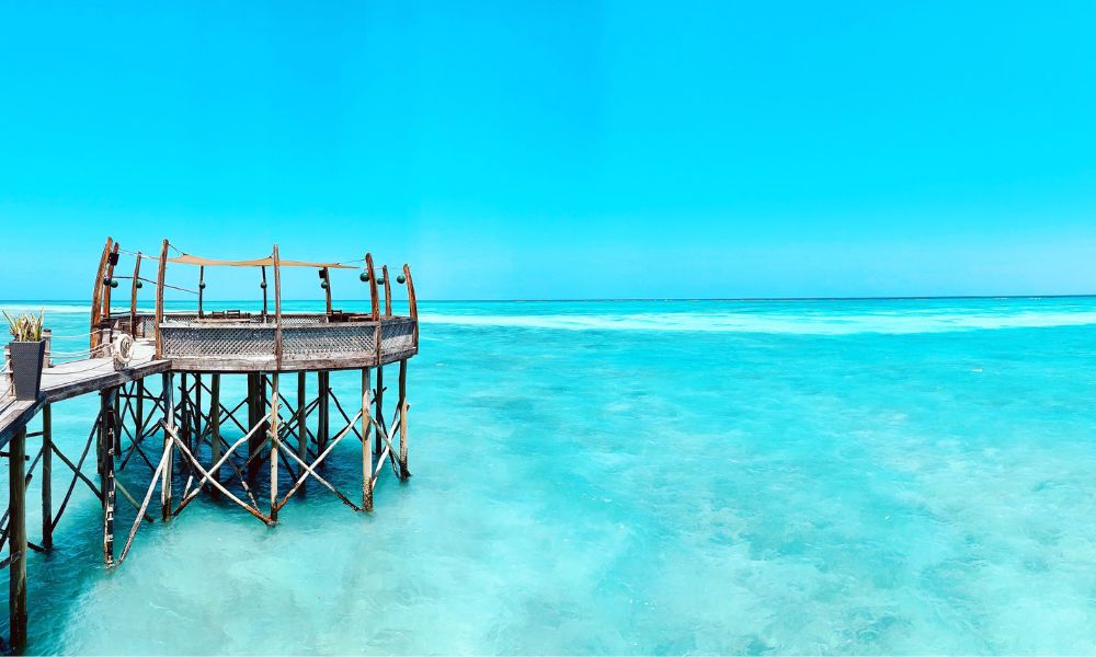 A turquoise sky meets sparkling turquoise waters. There's not one single cloud in the sky. And a wooden jetty/bridge is extended out in the waters - this is Zanzibar in Tanzania. 