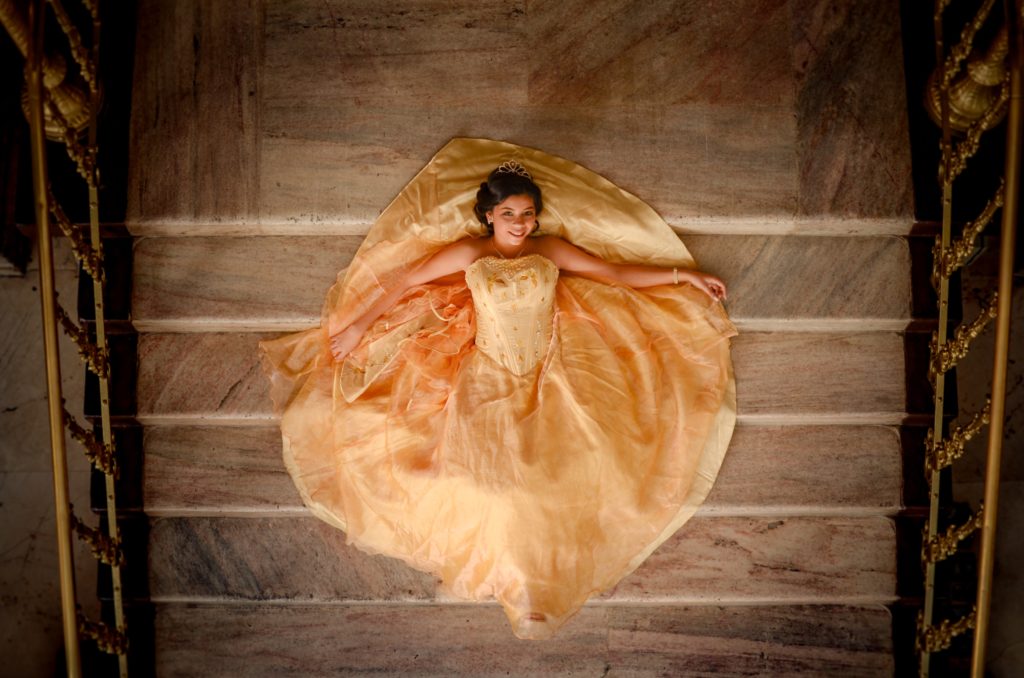 A woman dressed in an orange gown and a tiara lies on the stairs of an ornate mansion - she is smiling and the shot is taken from above