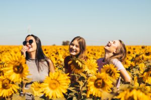 Three women are in a sunflower field. They are laughing, smiling - one of them has her head back in laughter and all the sunflowers are blooming. Three close friends on perhaps a bestie break of sorts.