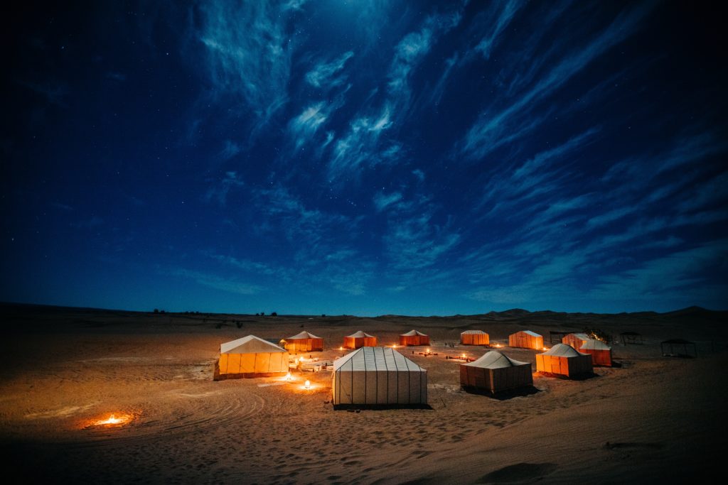 A canopy of  a sparkling blue sky hovers over tents laid out in the desert. You can also see the glow of several small campfires.