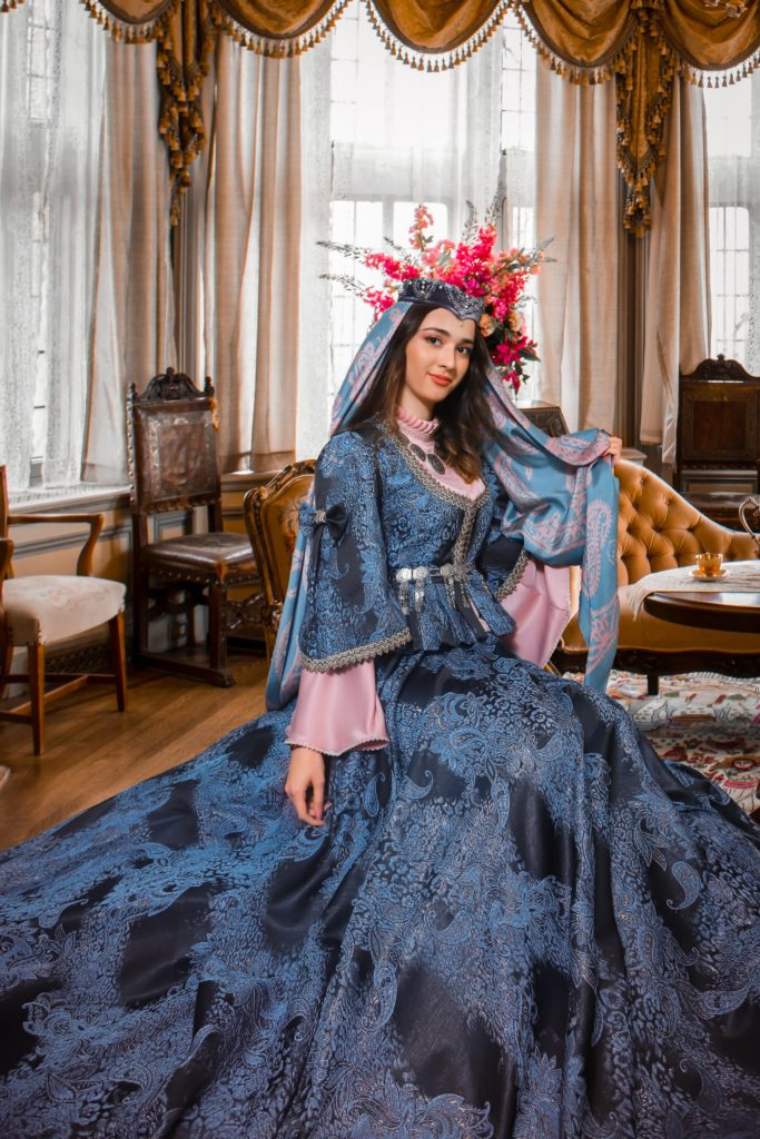 A woman wears a blue gown like dress and is sitting in an ornate room, smiling at the camera. It is a period costume of some kind and the material looks like satin