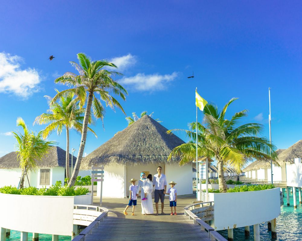 You can see a luxury hut kind of a location with blue skies, palm leaves, white beach sand, and a happy family. They look like they are on an all-inclusive holiday or an all-inclusive resort somewhere in the Maldives