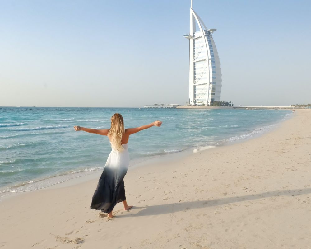 A woman on a Dubai beach seems to seek the winter sun - you can see her in a white and black summer dress, arms outstretched as she is walking on a beach and in the horizon you can see a landmark building of Dubai