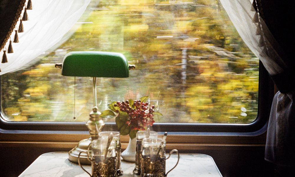 A view from a window seat/s in a train journey - the window has white and brown curtains with tassels, and there is a lamp, a floral arrangement and two vintage cups with spoons on the table between the seats 