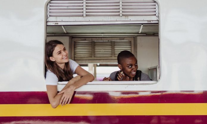 Two young women stare out of a train window Both are smiling and seem happy with their train journey. One woman is white or Caucasian and another is black