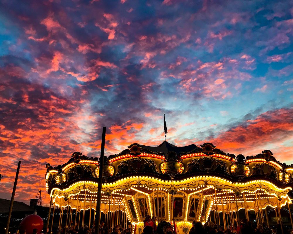 A crimson sky is spread over a brightly lit carousel ride in an amusement park. It is nighttime and you can make out that the theme park is crowded - it could be a weekend or mid-term break