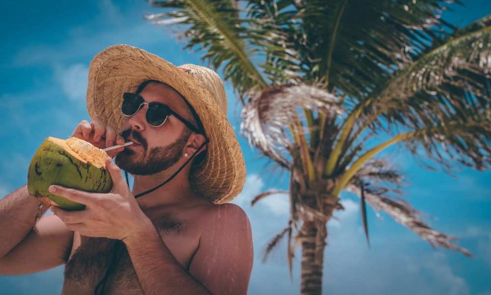 A man on a vacation with a straw hat and sunshades sips on a fresh coconut and in the background are blue skies with a palm tree 