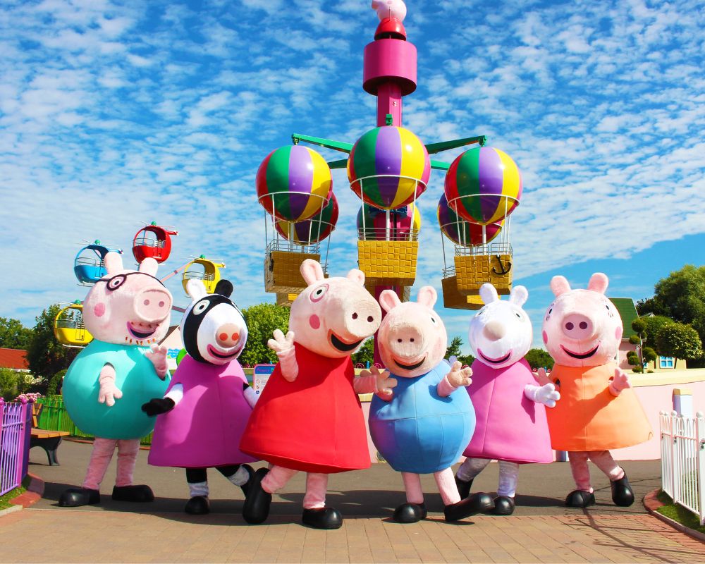 You see various five Peppa Pig characters in their distinctive costumes and colours and behind them you can see an amusement ride and blue open skies. This is from Paultons Park and makes for a good mid term break for children