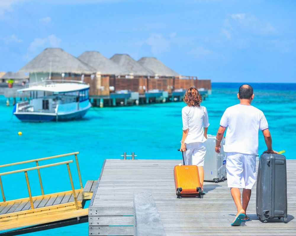 A man and a woman with suitcases head to huts (looks like a beach resort). The water is blue and so are skies, and the man and woman are wearing pristine whites. It seems like an all-inclusive holiday that couples book for anniversaries