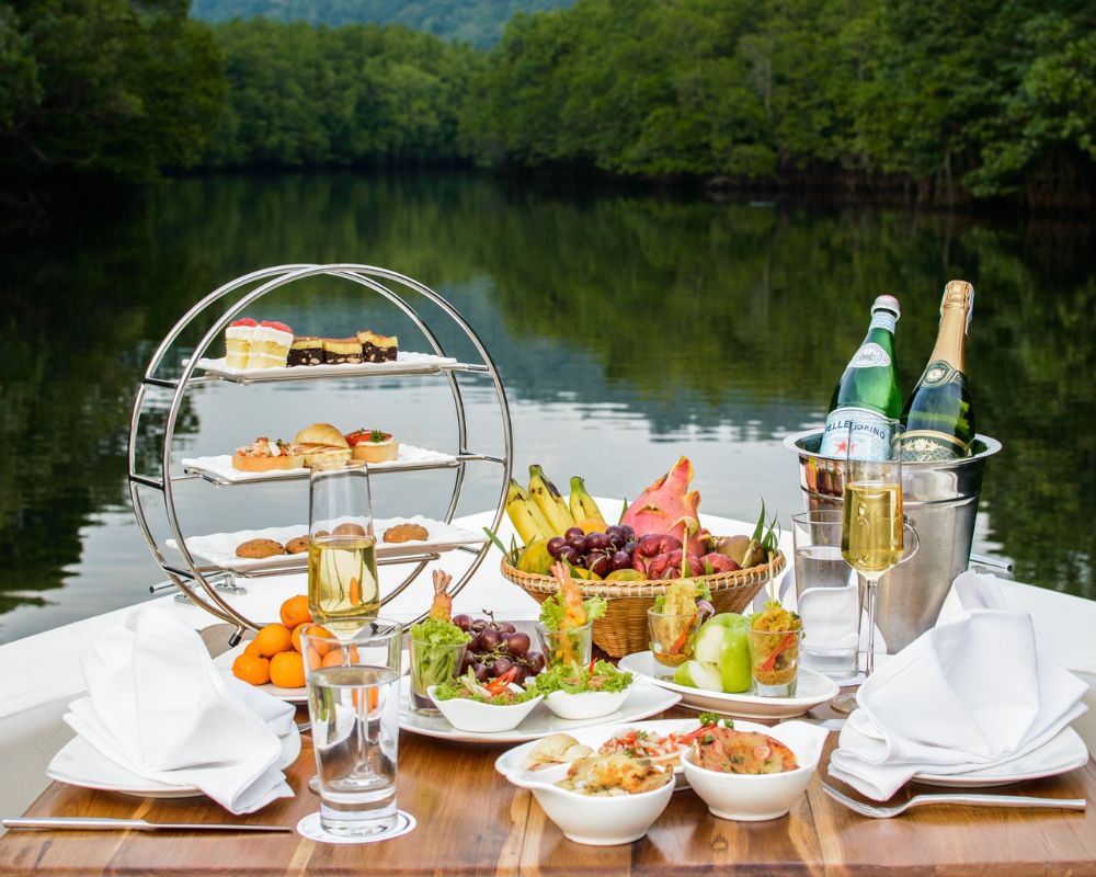 A photograph shows food laid out facing the waters. It has a stand which has pastries, biscuits and sweet treats. There is a bowl with various fruits and a bucket of drinks and glasses filled with drinks and white napkins.  Also food in bowls and serving platters 