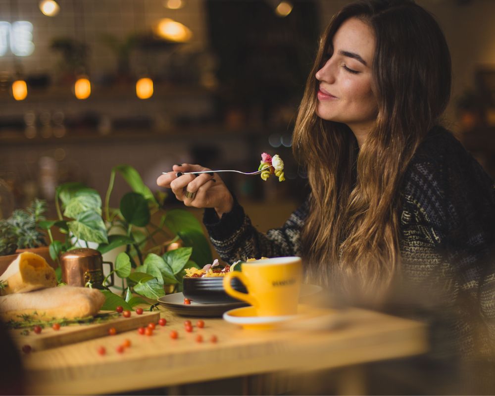 A woman sits inside a café. Her eyes are closed as she savours the fragrance of a meal. There is a coffee cup on the table and she seems to enjoying like a local in a new city