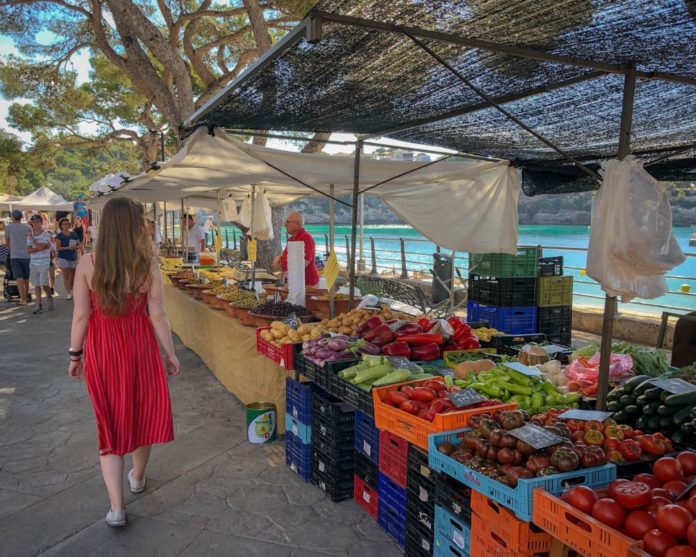 A woman in a red dress, and hair till her waist walks past a local market that is full of fresh produce