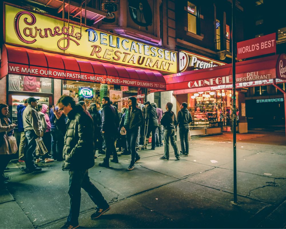 It's night time and locals are queueing up at a place called Carnegie Delicatessen Restaurant. A line beneath the billboard's main slogan says 'we make our own pastries' and follows it with meats, pickles, prompt deliveries,