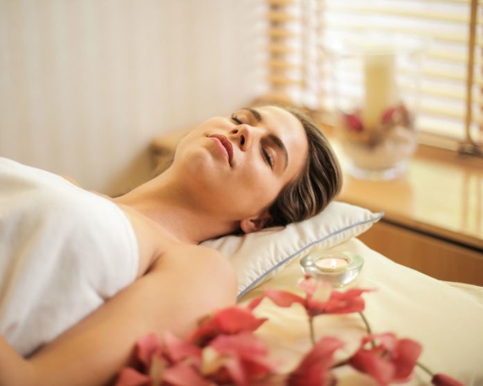 A woman sleeps on a bed - she is having a spa or a massage experience. Rose petals and flowers are visible as also a scented candle
