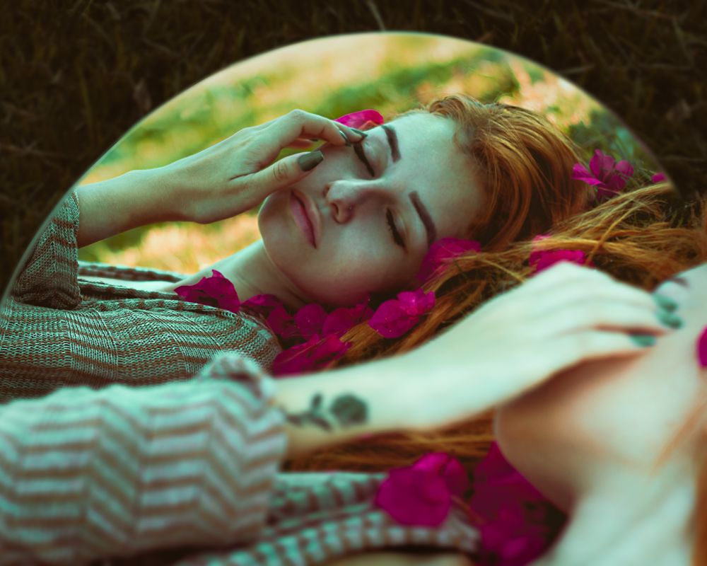 A woman with a tattoo on her wrist and orange hair sleeps, her hand is on her face and there is a mirror next to her so that you can see her reflection. There are flowers around here - perhaps she is enjoying a spa like moment or a package offered under sleep tourism.