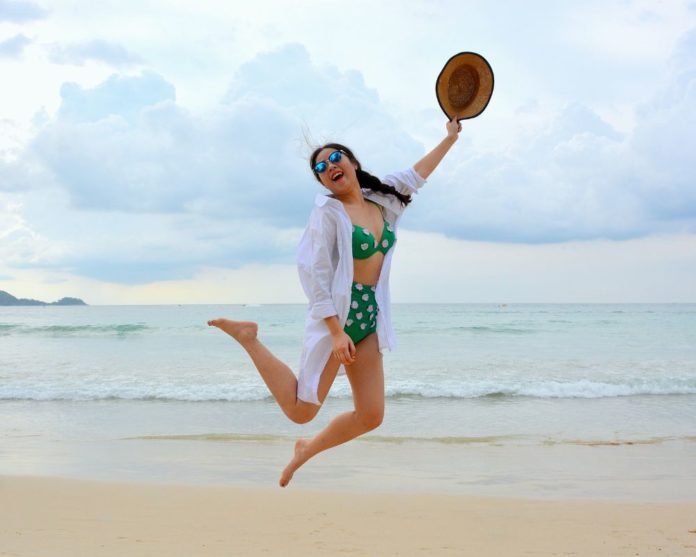 A woman in a bikini and a hat in her hand jumps up in the air on a beach in summer