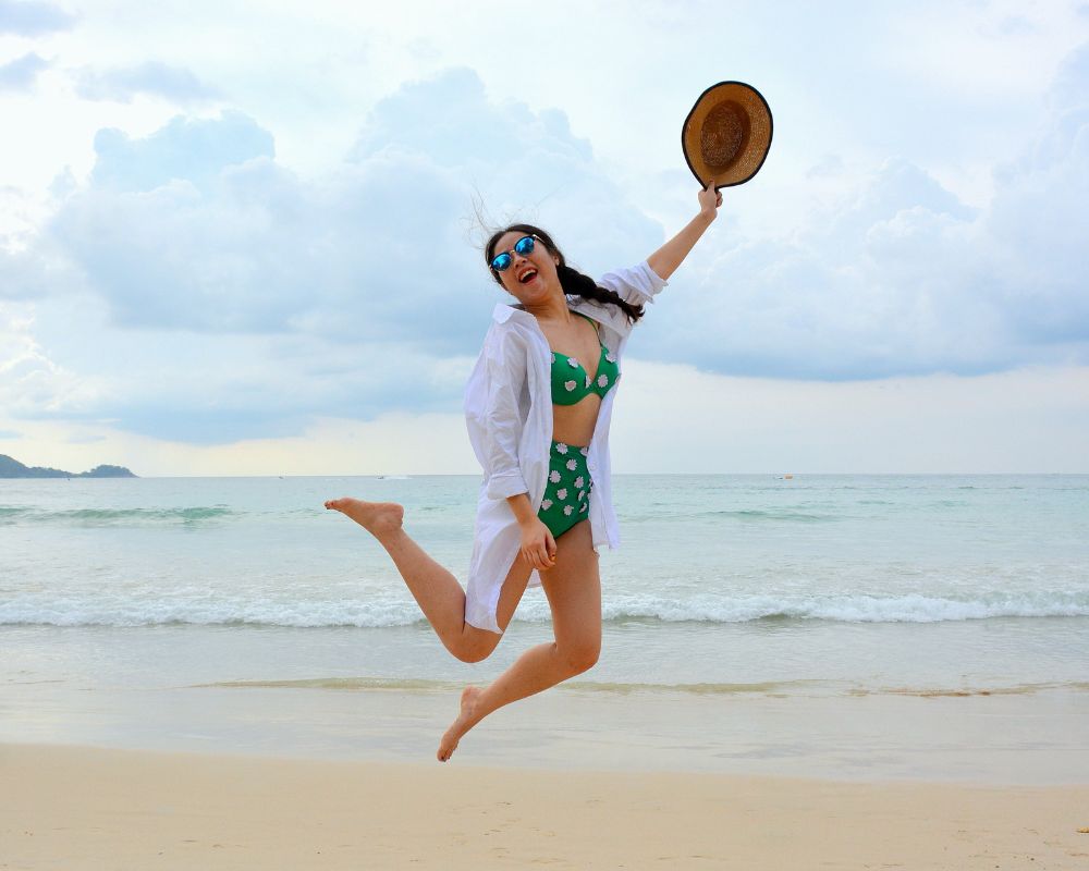 A woman with black hair in plaits, and a green bikini and a white shirt over it leaps up in the sky on a beach. She holds a straw hat in her hand and has sun glasses on. She looks happy and radiant as if this holiday has been good for her and her overall wellbeing 