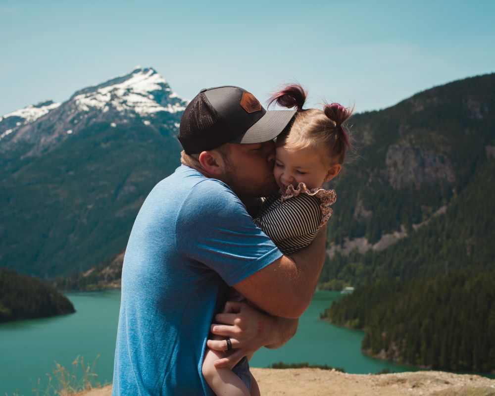 A man, a person of colour (black) is wearing a cap and holds his toddler daughter close to him, giving  her a kiss. She is in his arms, they are happy, on a travel trip and behind them you see mountains and a lake