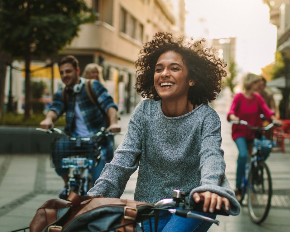A woman with curly hair on a bicycle and two more people are behind her. The woman is smiling and she is a person of colour. 