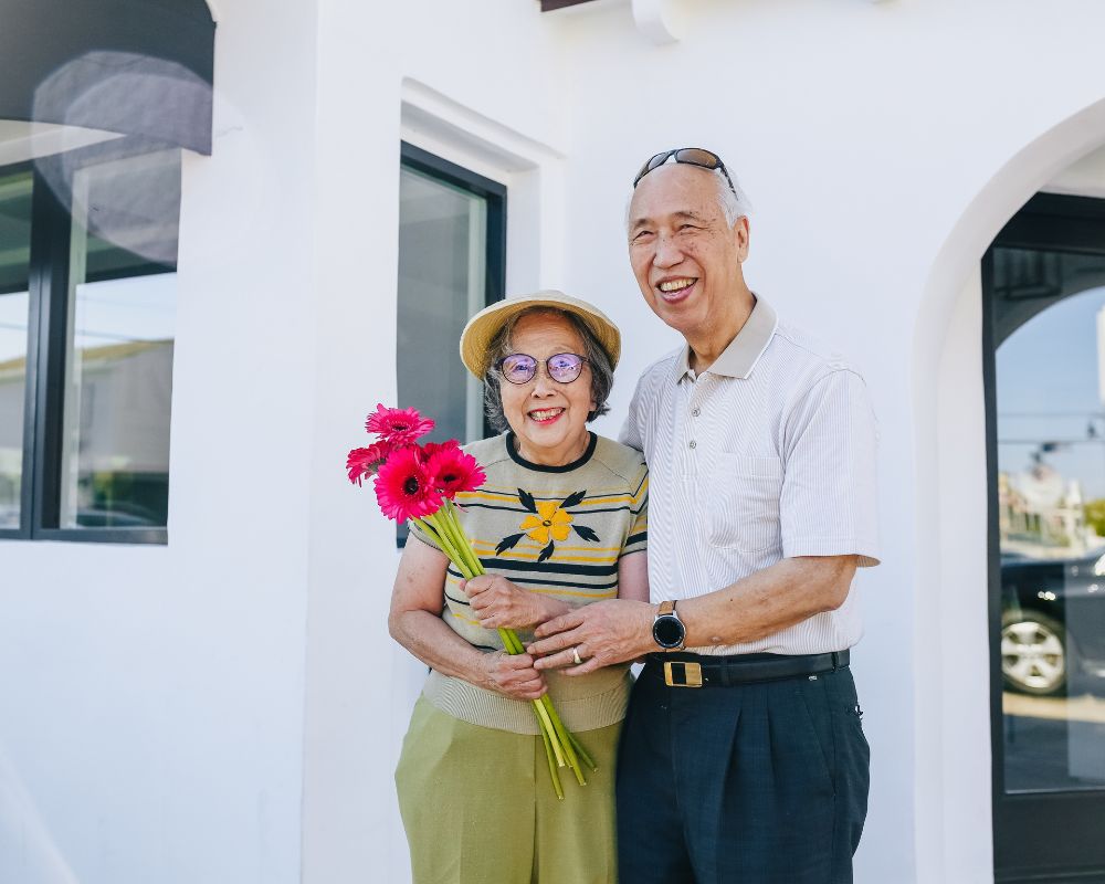 A man and woman - they seem to be in their sixties - stand outside a building. The woman is holding flowers, the man holds her hands and they are smiling. They seem like they are out on a holiday, making time for themselves and for their wellbeing 
