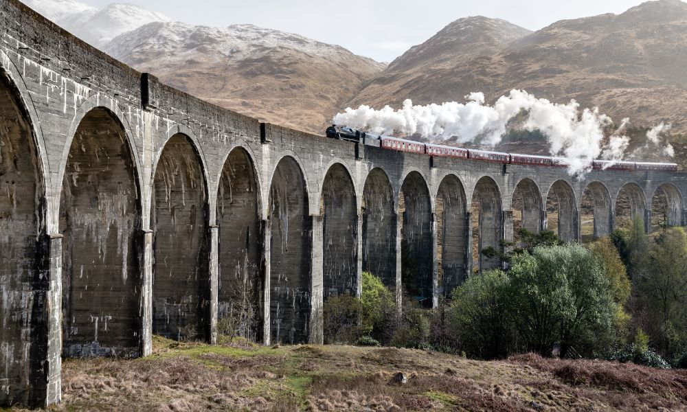 You can see a train with puffs of smoke going over a via duct. In the background are hills and mountains. It seems like passengers are on a very scenic train journey. 