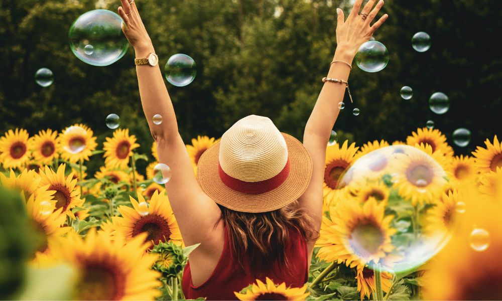 A woman in a field of sunflowers and very she has her hands up and is wearing a hat