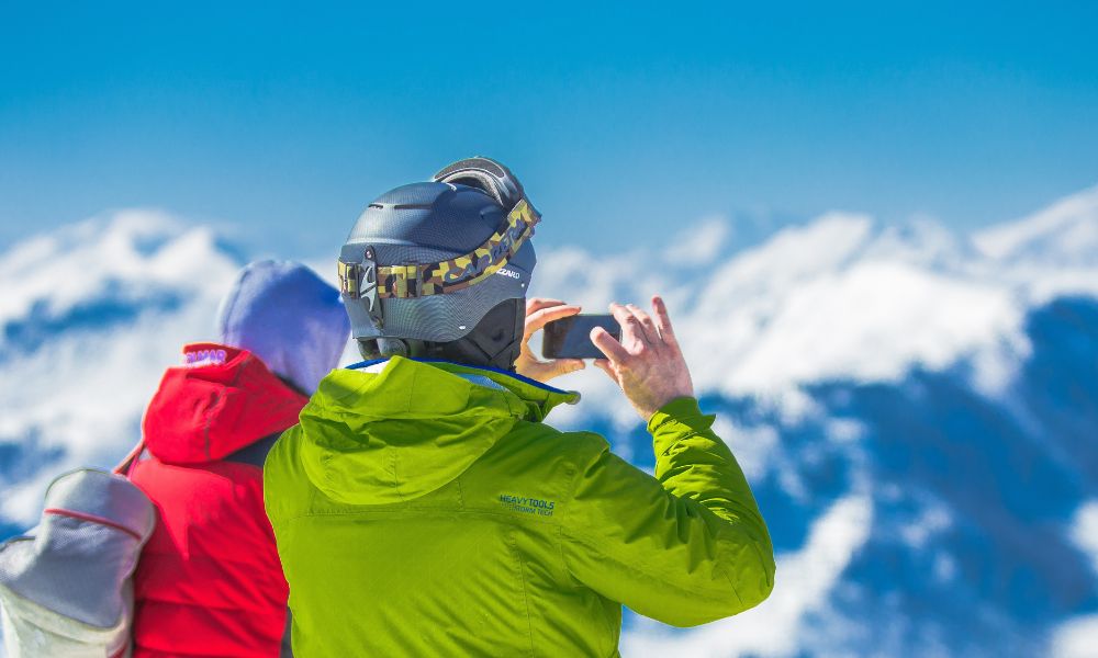 A man in green and a woman in red (you see their backs) are in ski gear and taking a photograph of snowy mountains - perhaps a pic that will raise holiday envy among their friends!