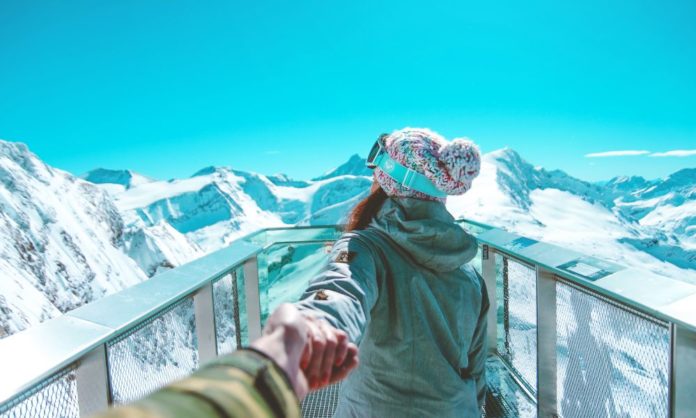 A woman holds a man's hand (you can't see their faces) and seems to the pose that says come with me to a spa holiday. In the background are snow capped mountains and they are on a ski terrace