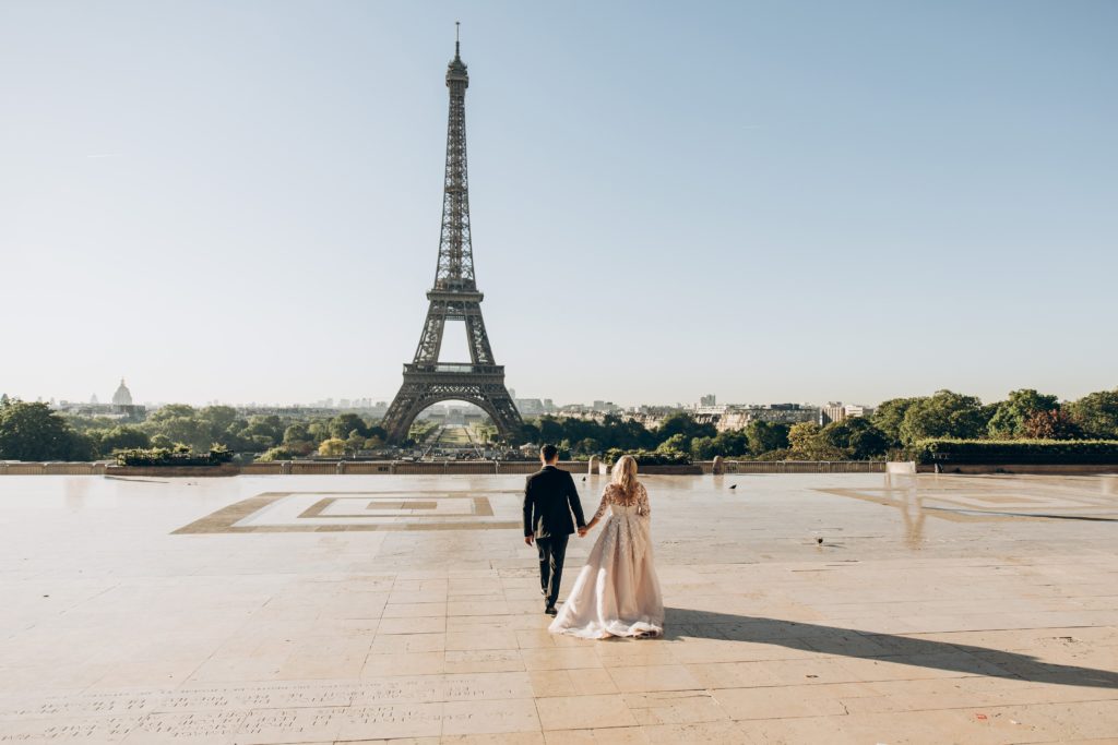 A man and a woman with their backs to the Eiffel Tower in a romantic pose - holding hands. Man is in a suit, and the woman is in a white dress.