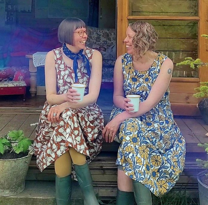 Two women sit together in beautiful print dresses, holding a cup each and smiling at each other
